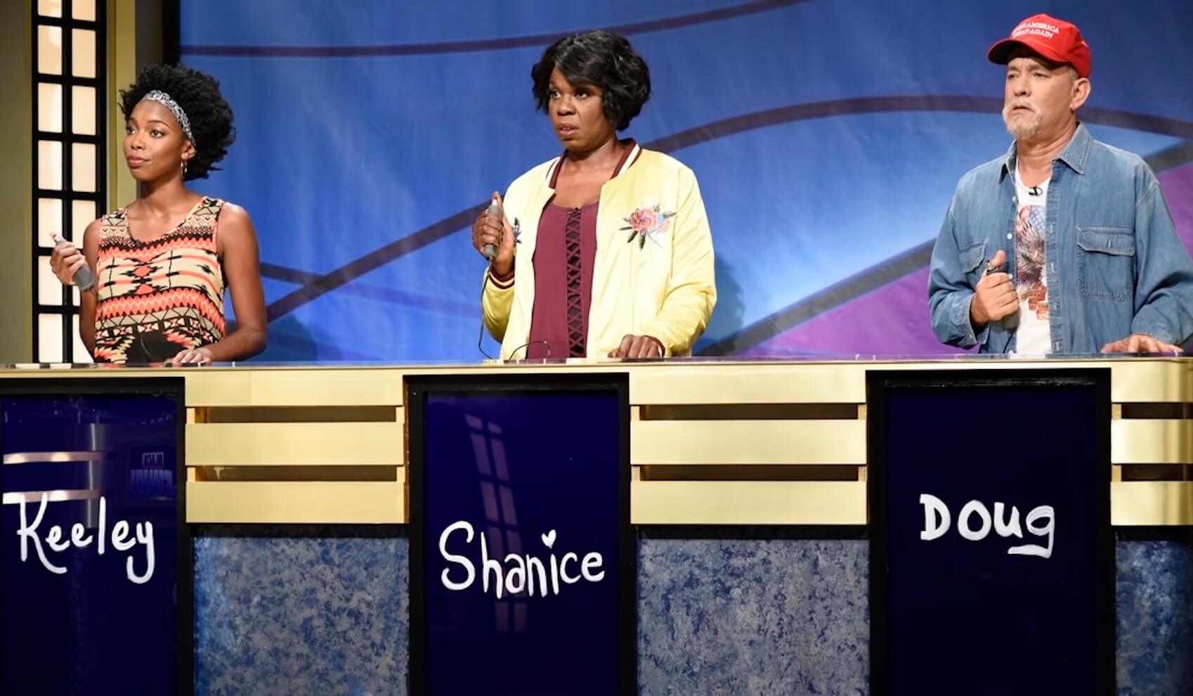 Two black women and a white man with a red hat on playing a game show during a skit for 'Saturday Night Live'
