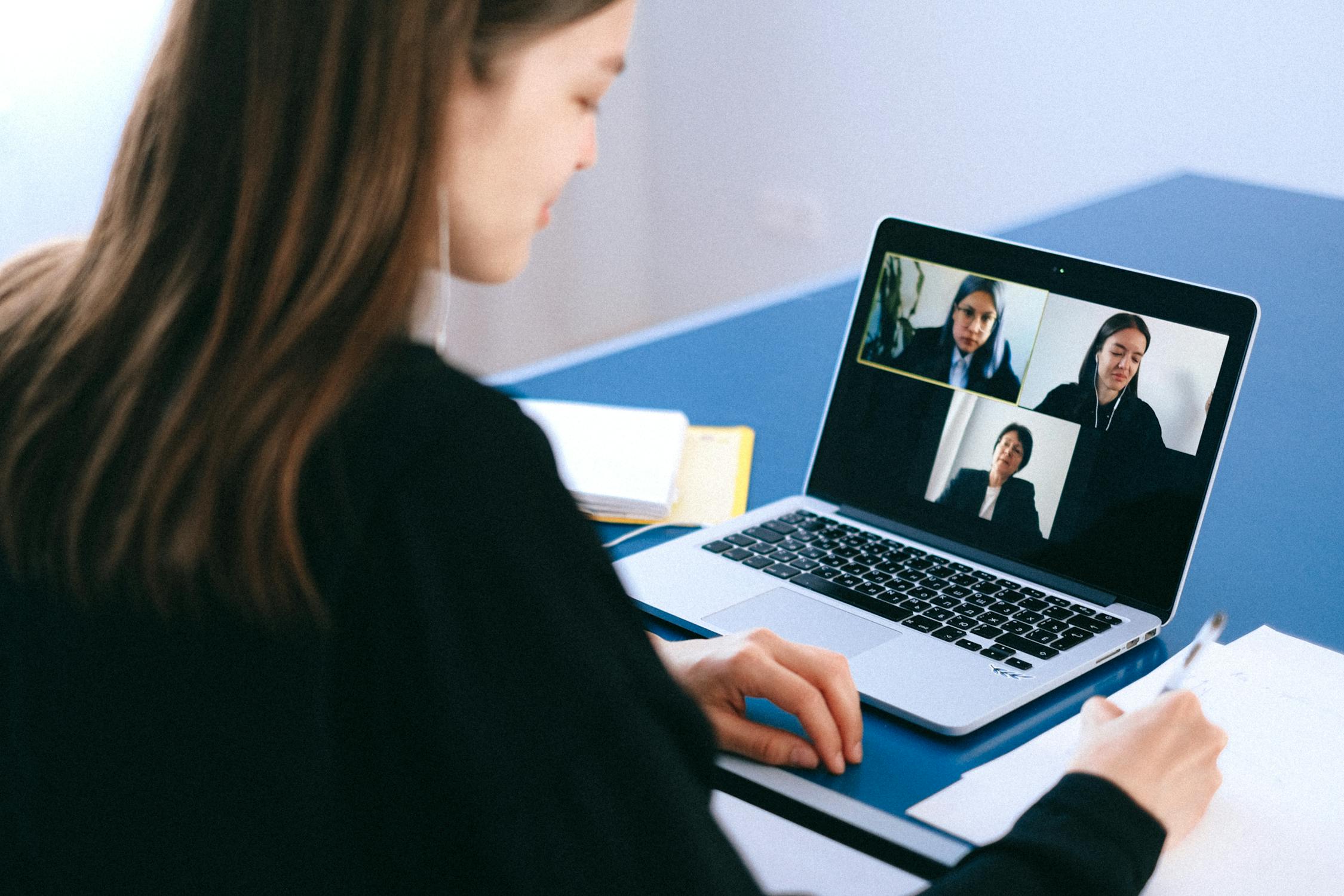 A woman with long brown hair writing as she attends a Zoom meeting with two others